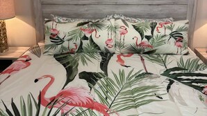 A bedroom with a bed featuring a floral and flamingo print, a wooden headboard, and a painting of flamingos and flowers on the wall.