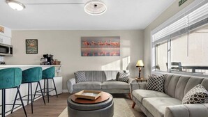 A modern living room with a grey sofa, teal bar stools, a coffee table, and a kitchen area with a microwave and a painting of flamingos.