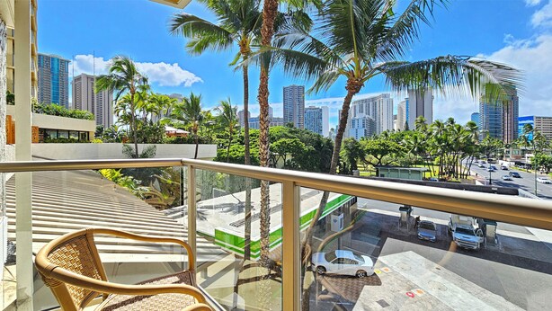 A balcony with a wicker chair, overlooking a cityscape with palm trees and high-rise buildings. - Luana Waikiki Hotel & Suites 119-Modern Waikiki Studio,2 Beds,AC,WiFi,Pool & Gym (Honolulu)