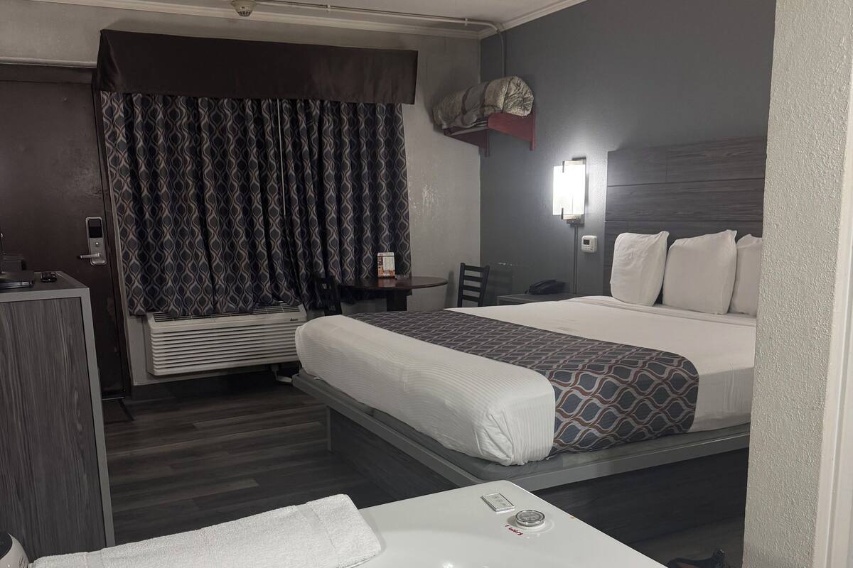 A hotel room with a large white bathtub, two beds with patterned bedding, a desk with a chair, and a window with curtains.