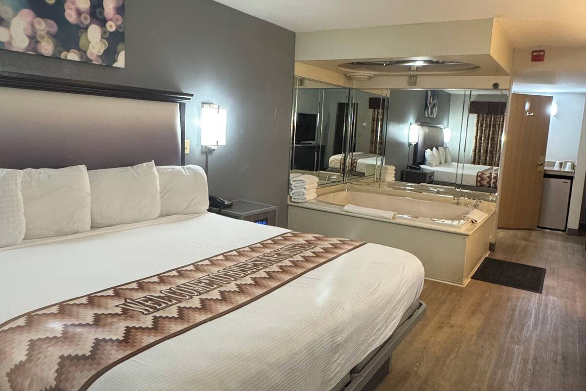 A hotel room with a large bed, a glass-enclosed bathroom, and a mirror reflecting the room.
