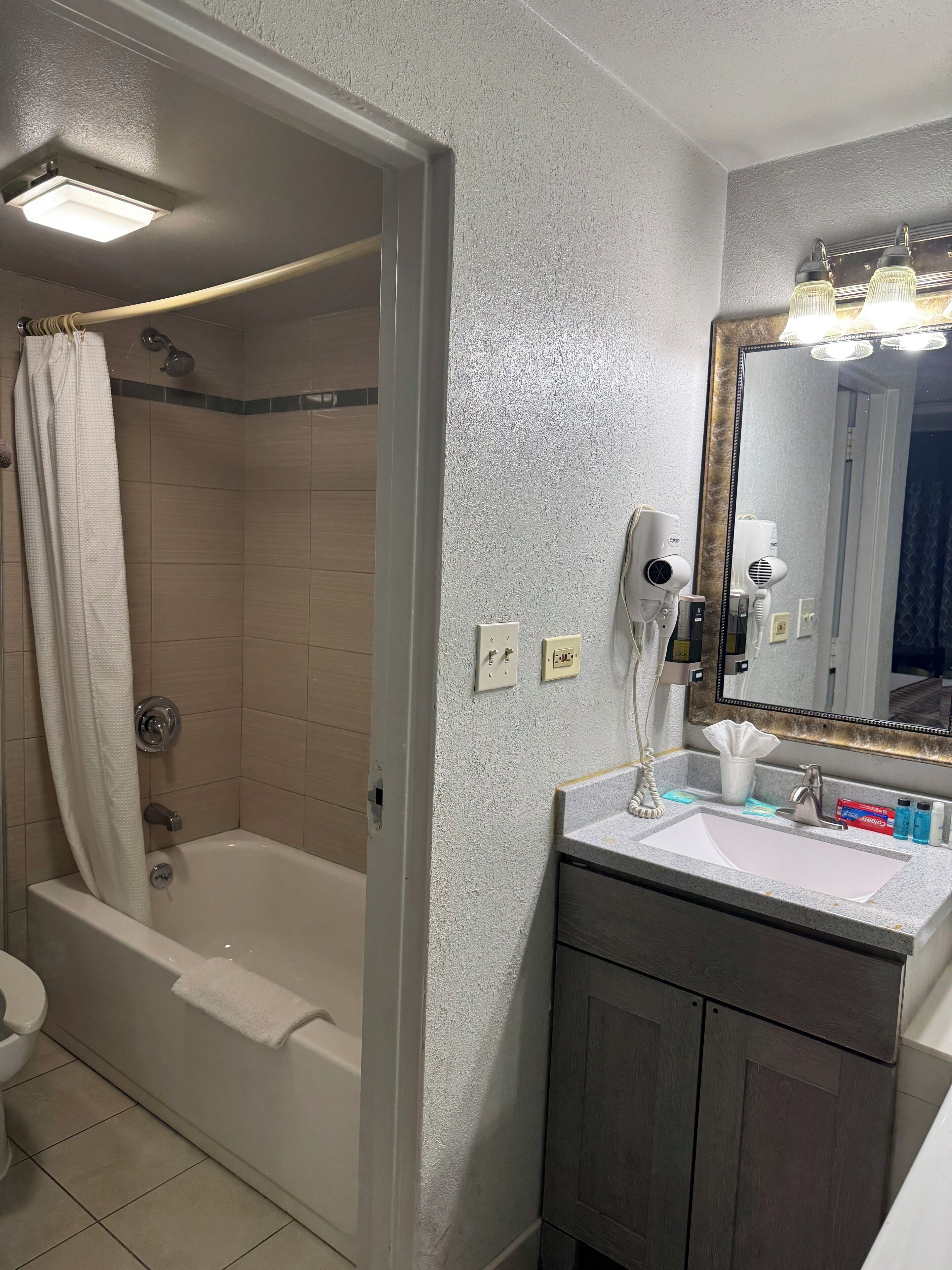 A bathroom with a bathtub, a shower, a sink with toiletries, and a mirror with lights.