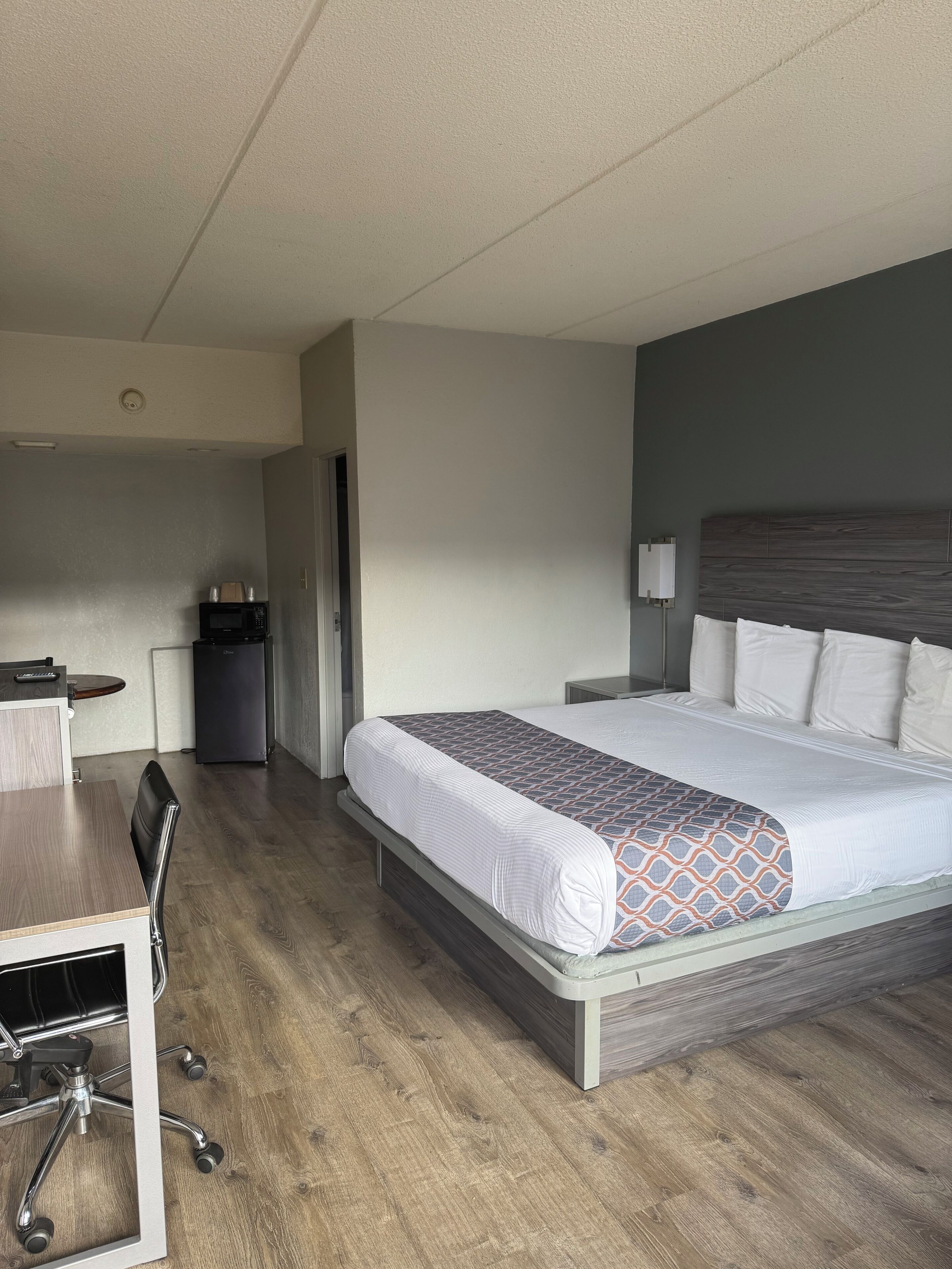 A hotel room with a bed, a desk, a chair, a microwave, and a trash bin.