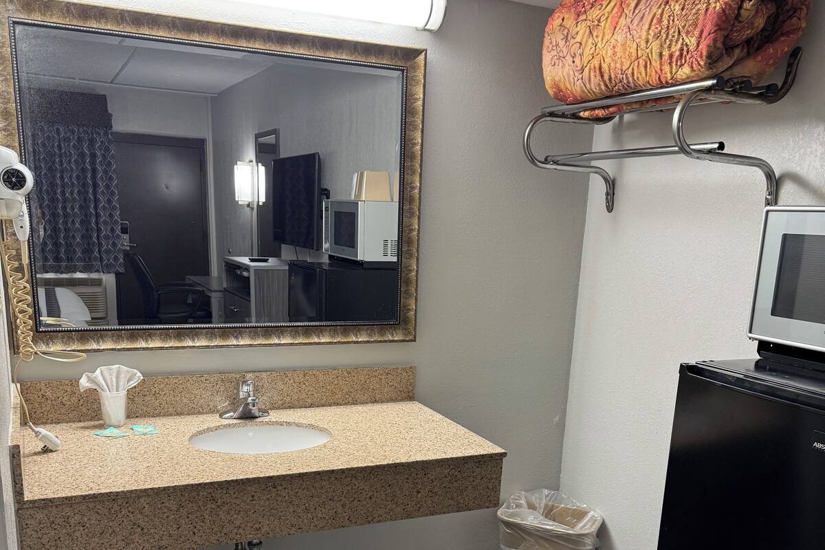 A bathroom with a granite countertop sink, a large mirror, a hairdryer, a microwave, and a black refrigerator.