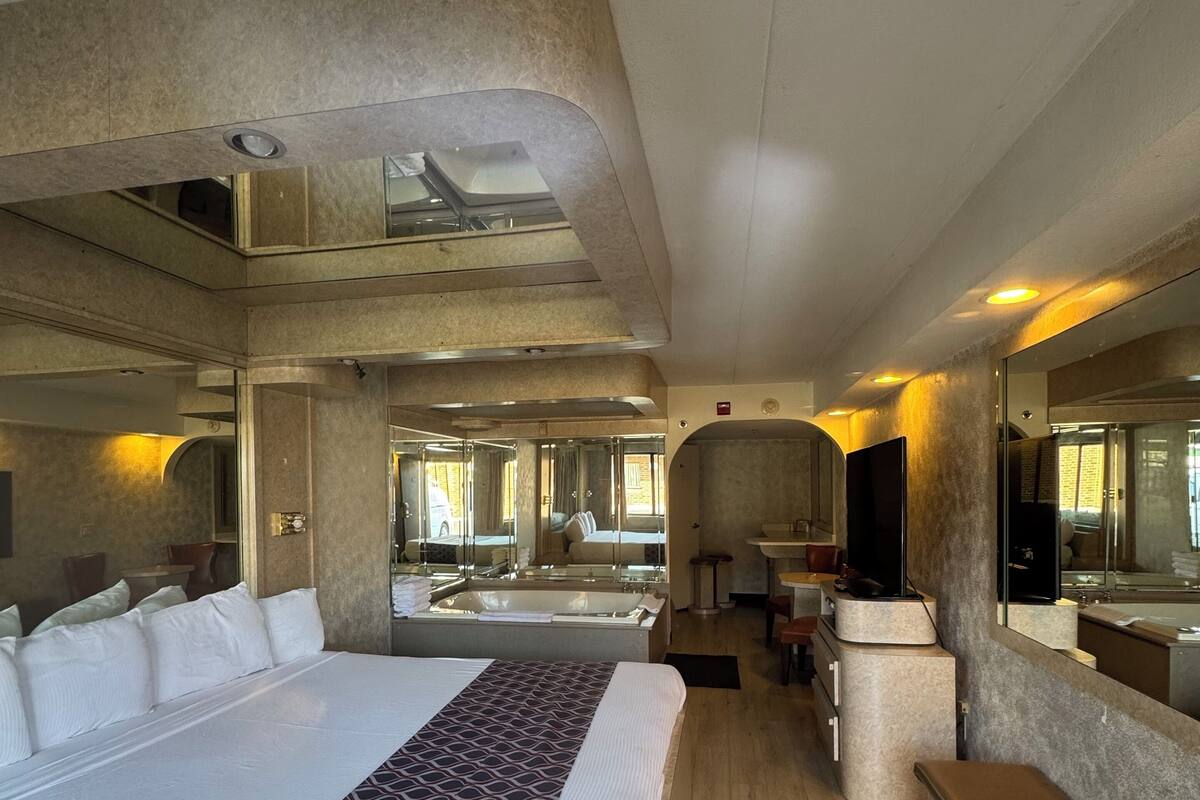 A modern hotel room with a large bed, a sitting area, a bathtub, and a large mirror.