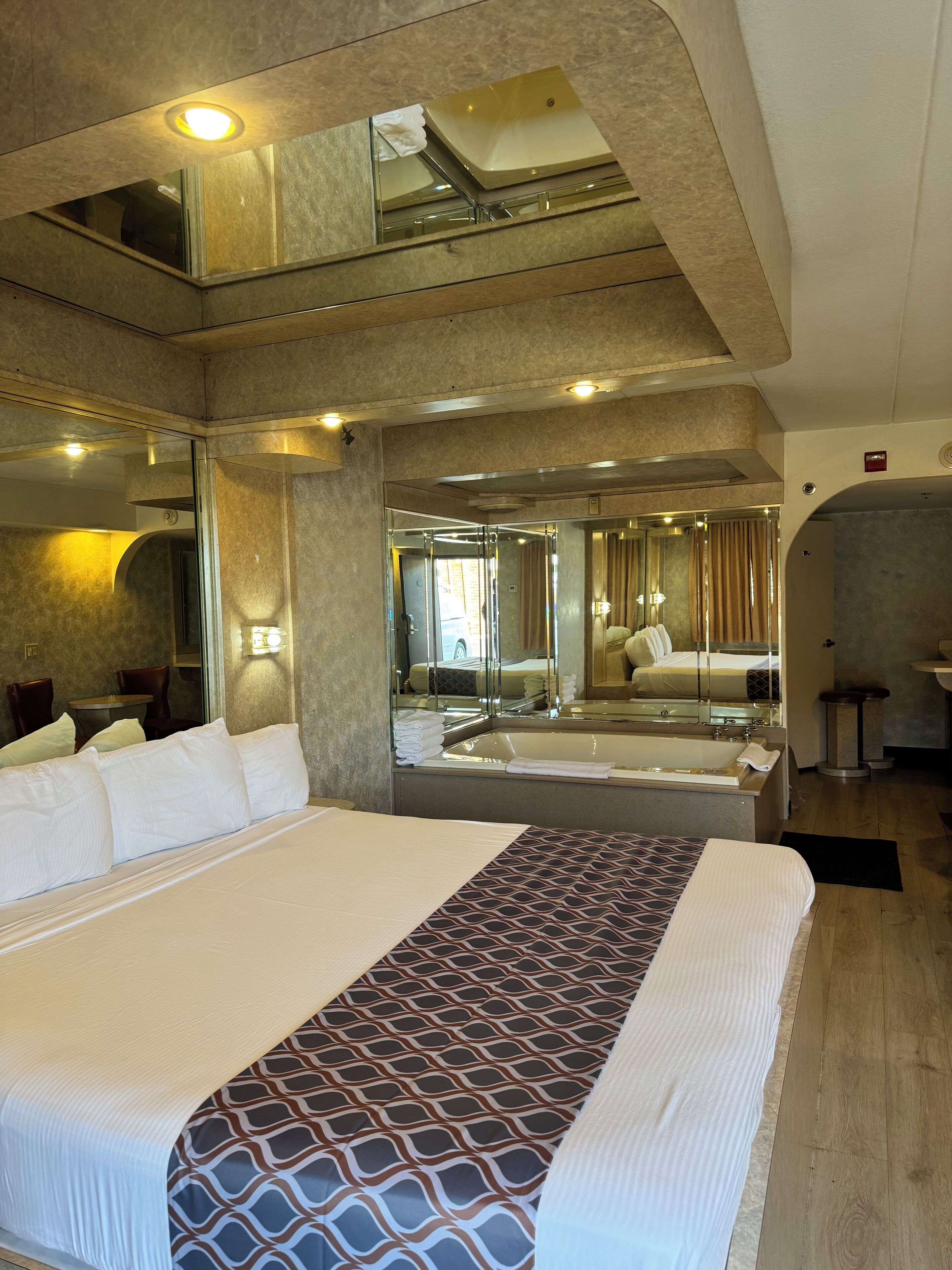 Une chambre d’hôtel avec un grand lit, une salle de bain avec une baignoire et un miroir, et une petite table avec des chaises.