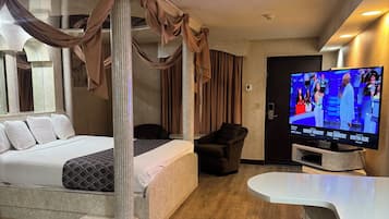 A modern hotel room with a four-poster bed, a TV, a bar area, and a seating space.