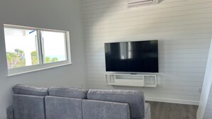 A modern living room with a grey sofa, a mounted flat-screen TV, and a window with a view of greenery.