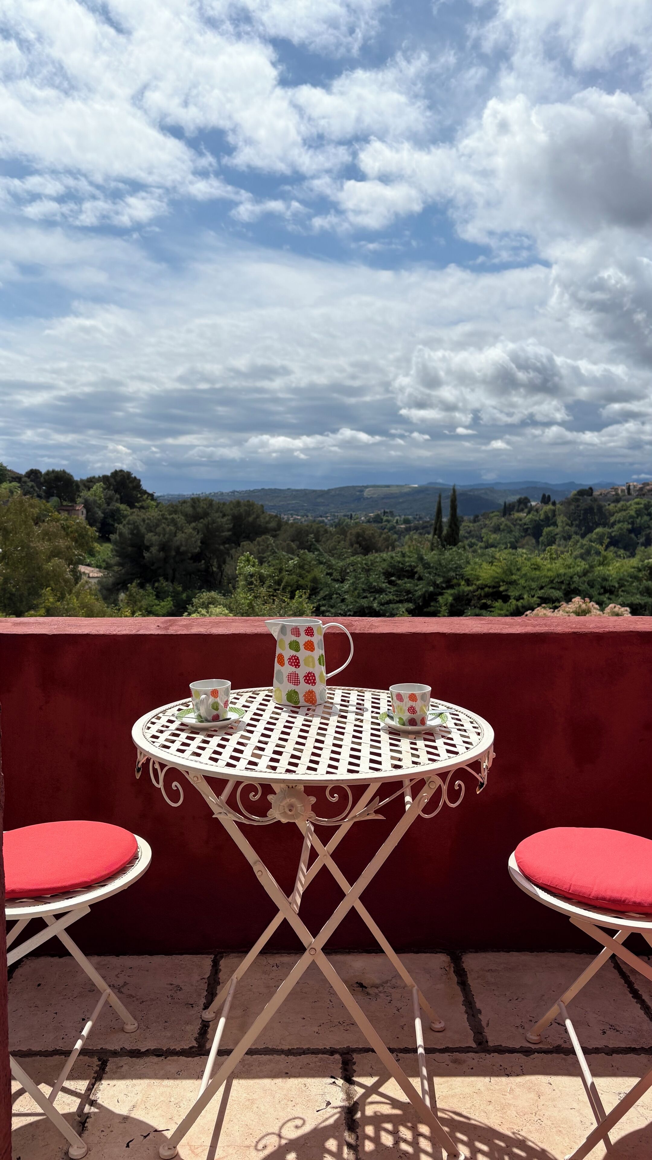 Mesa metálica redonda con cojín rojo, una jarra y dos tazas en un balcón con vistas a un paisaje pintoresco.