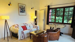 Een gezellige woonkamer met een bank, fauteuils, een salontafel en een tafelvoetbaltafel. Er is een raam met uitzicht op groen.
