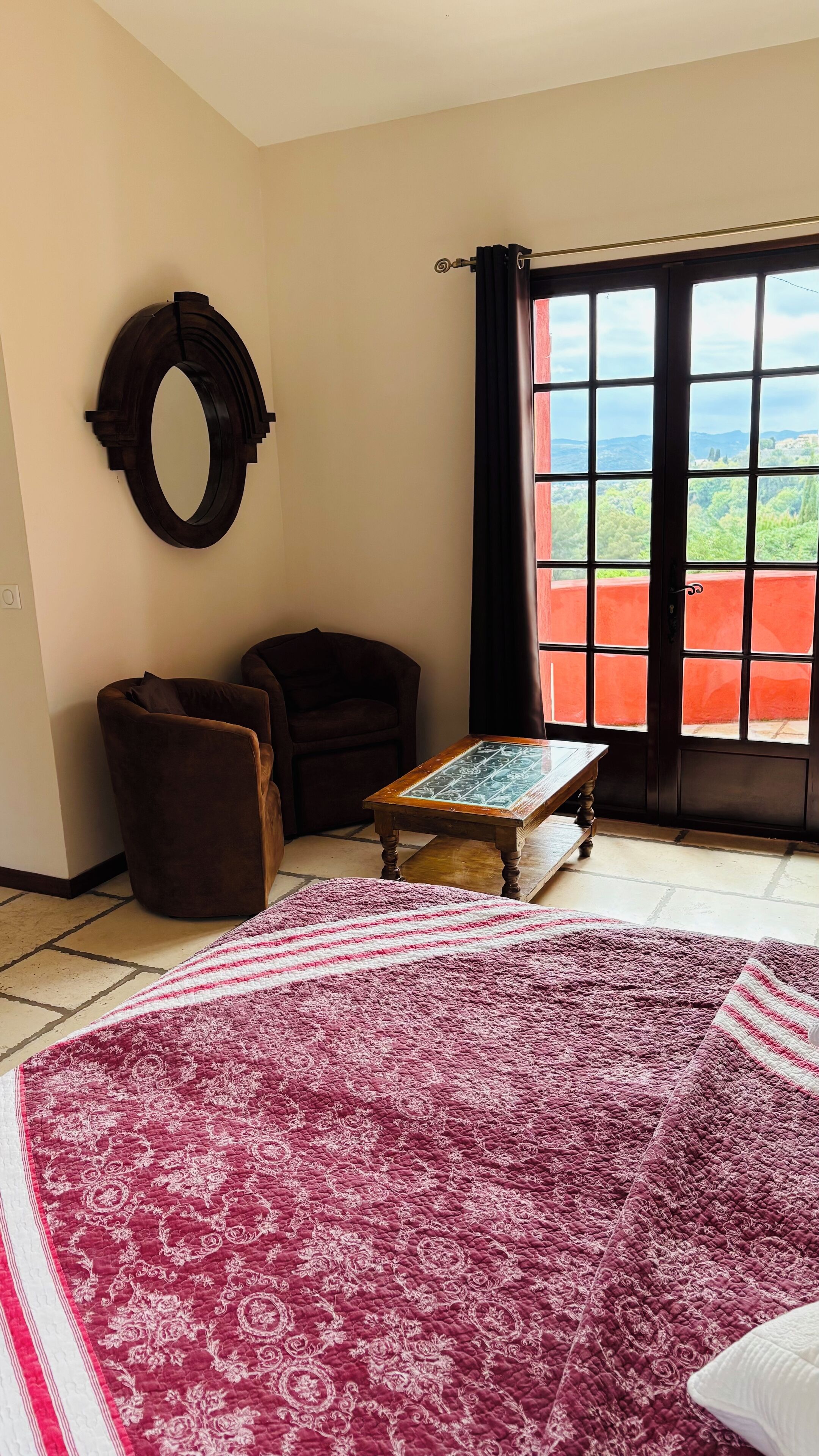 A room with a large window, a round mirror, a brown armchair, a wooden table, and a patterned rug.