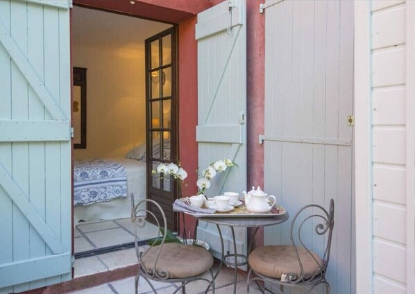 Ein kleiner Sitzbereich im Freien mit Tisch und zwei Stühlen, eine Tür, die zu einem Schlafzimmer führt, und ein Fenster mit weißen Blumen.