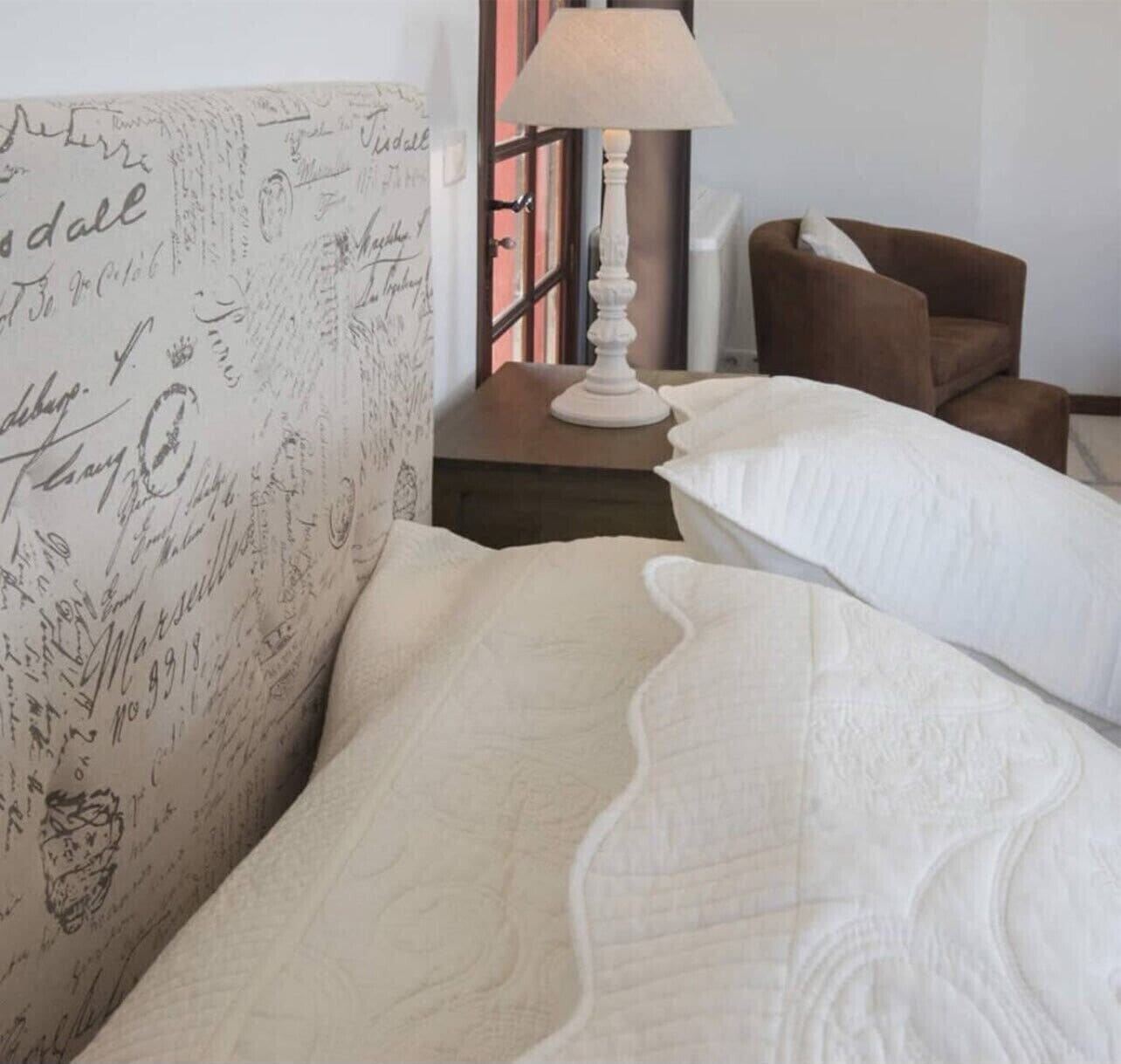 A neatly made bed with white bedding, a lamp on a bedside table, and a brown armchair.
