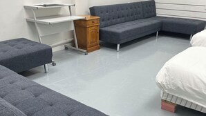 A modern, minimalistic room with a grey sectional sofa, a small wooden cabinet, and a white bed. - 3 single and 2 sofa beds Studio Suite 7 (Auckland)