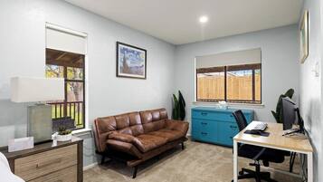 Een werkkamer met een bruine leren bank, een blauwe dressoir, een bureau met een computer en een raam met uitzicht op een houten schutting.