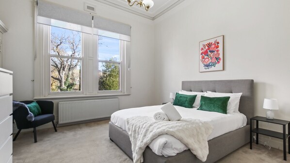 A bedroom with a large window, a bed with white bedding, a grey headboard, a black nightstand, and a chair with a green cushion.