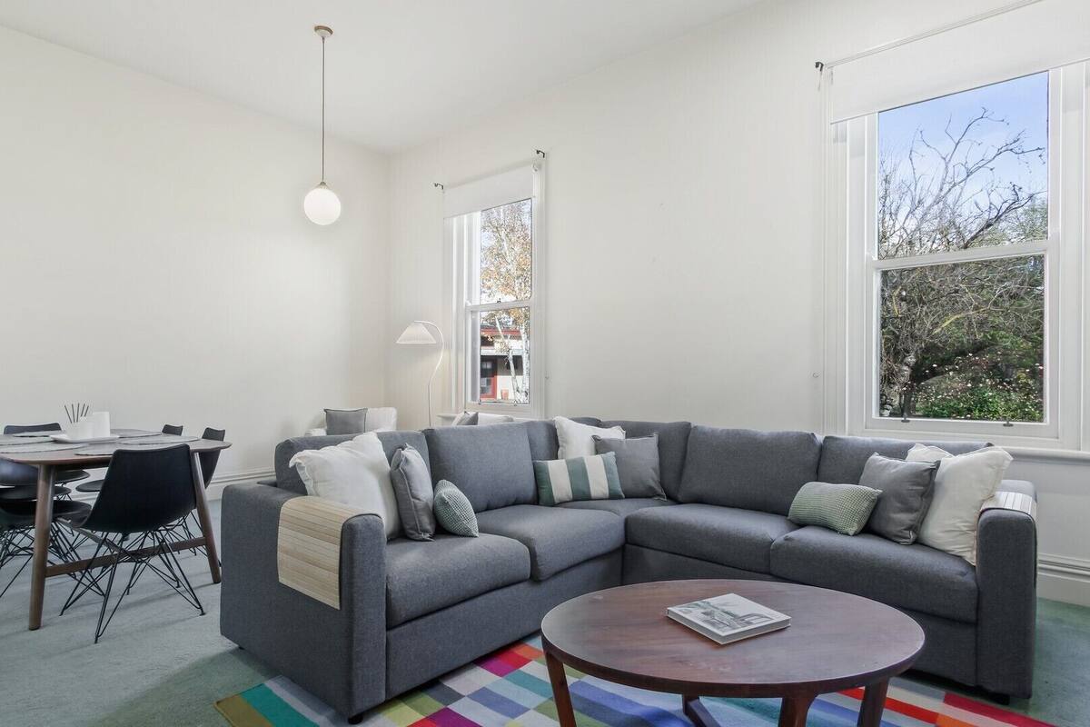 A modern living room with a grey sectional sofa, a round coffee table, a dining table with chairs, and large windows offering views of trees and buildings.