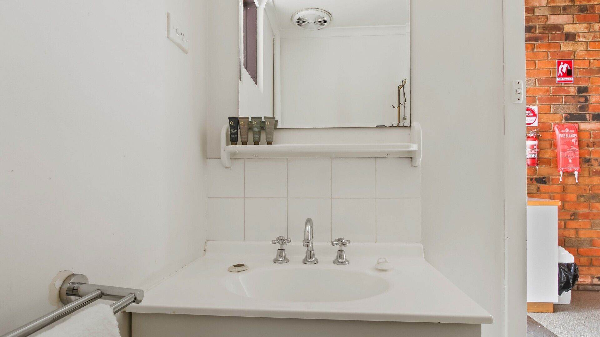 Un baño con un lavabo blanco, tres grifos, un espejo sobre el lavabo y un estante con artículos de aseo.