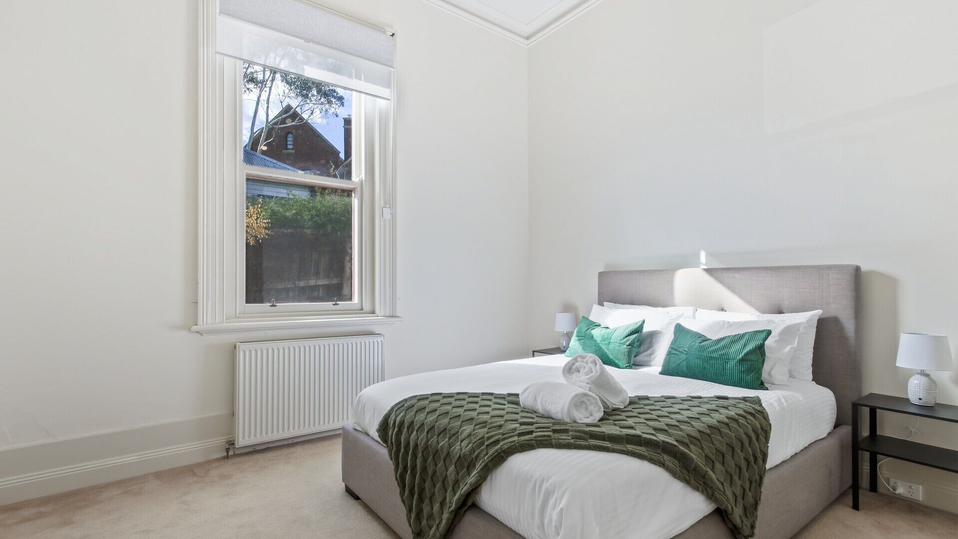 A bedroom with a large window, a bed with white and green bedding, and a radiator below the window.