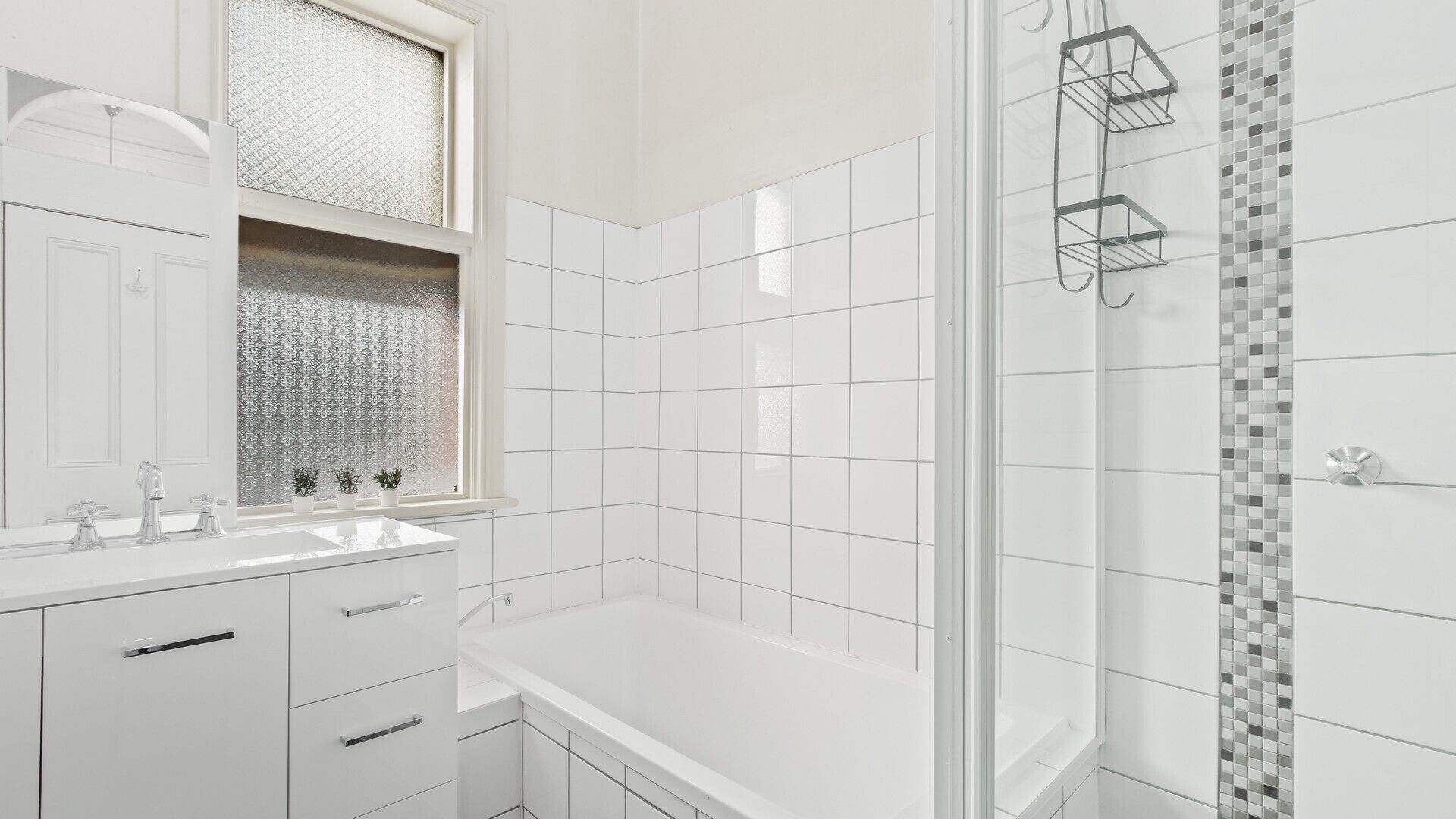 A modern bathroom with white tiles, a large mirror, a glass shower enclosure, and a countertop with a sink and three faucets.