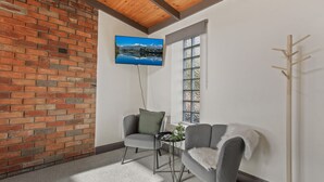 Una habitación con pared de ladrillo, un sofá gris, una mesita con una planta, un perchero de madera y un televisor montado en la pared.