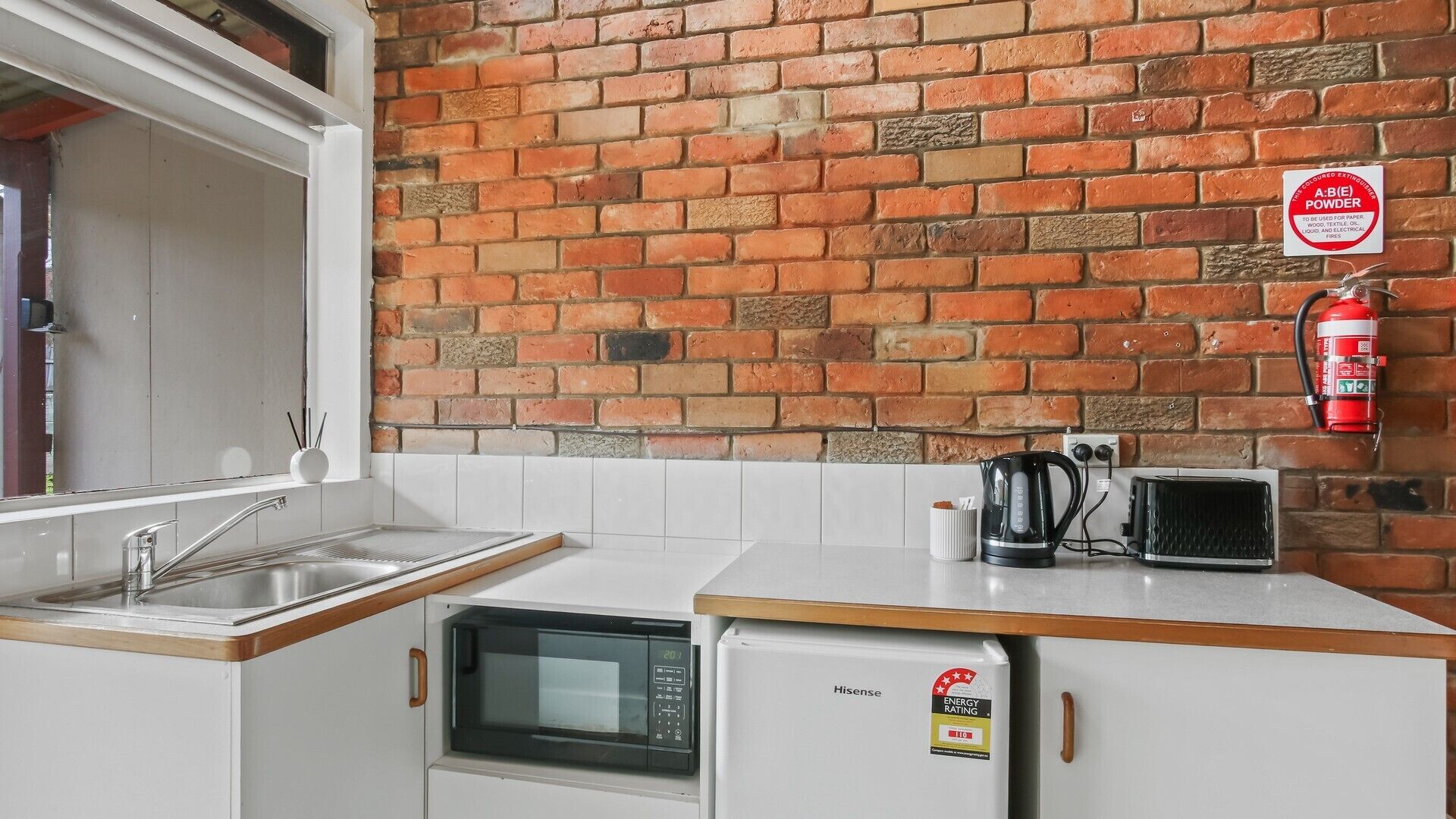 A kitchen with a brick wall, a sink, a microwave, a toaster, and a fire extinguisher.