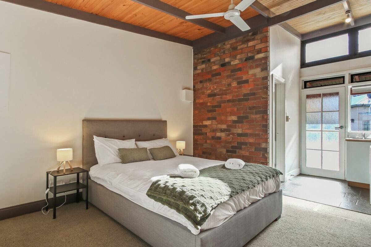A bedroom with a large bed, a brick wall, a ceiling fan, and a view of the outside.