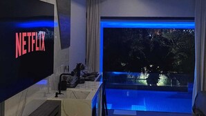 A modern hotel room with a large TV displaying 'NETFLIX', a fireplace, a balcony with a view of trees, and a bed. - Pousada Exclusive (Florianopolis)