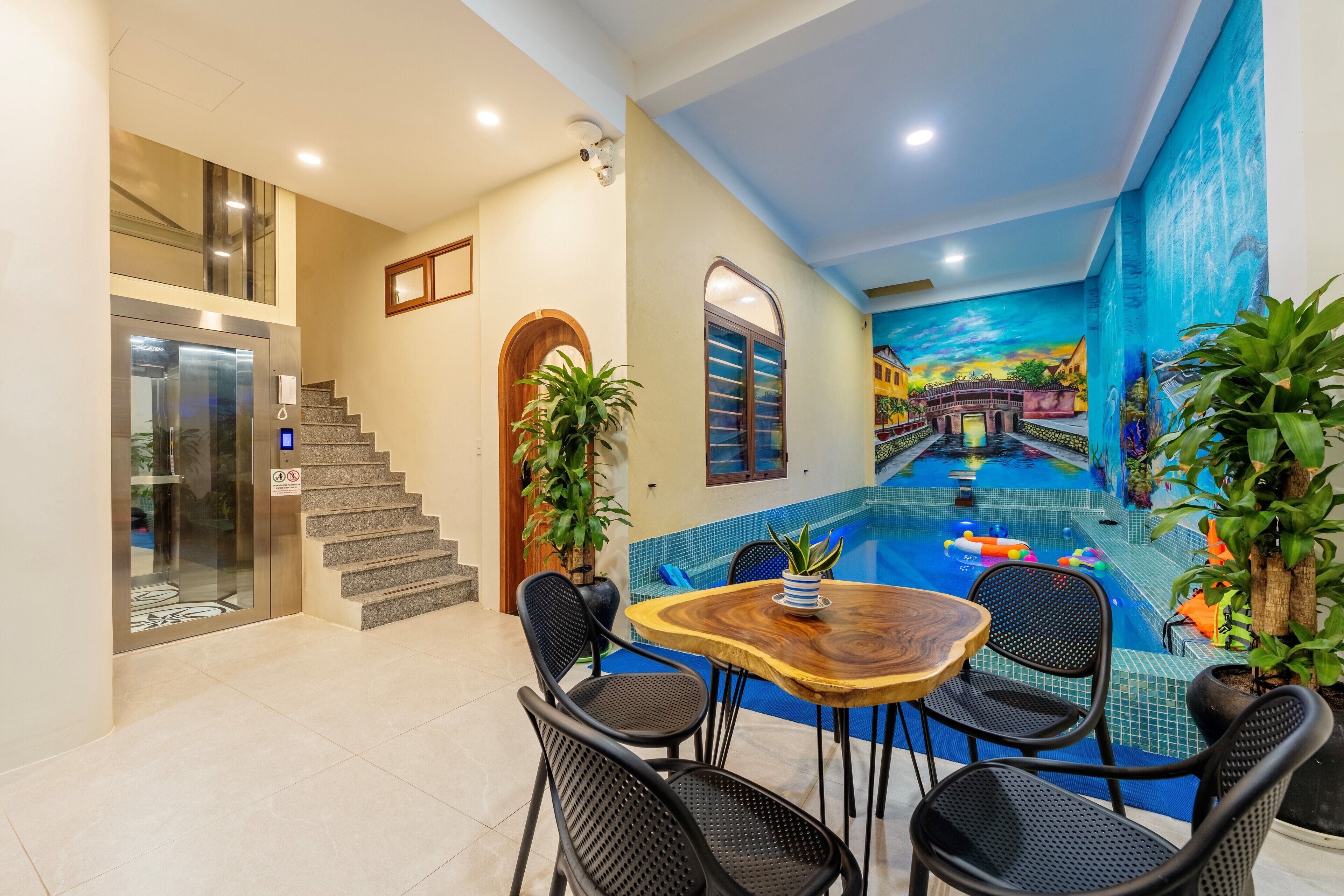 A modern indoor pool area with a wooden table and chairs, a staircase, and a mural of a tropical scene.