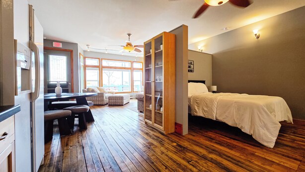 A hotel room with a wooden floor, a bed, a sofa, a dining area with a table and chairs, and a view of the outdoors through large windows. - 3 Min to Lake Michigan, 15 Min to Ski Crystal (X) (Elberta)