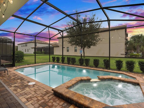 Piscina con jacuzzi, rodeada por un cerramiento de vidrio y vista a una zona residencial al atardecer.