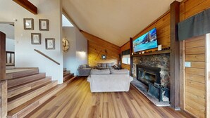 A cozy living room with a stone fireplace, wooden floors, a staircase, and a television mounted above the fireplace. - Close to Lifts | Townhome | Angel View 2-3 (Angel Fire)