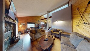 A cozy living room with wooden walls, a stone fireplace, and a large window. - Close to Lifts | Townhome | Angel View 2-3 (Angel Fire)