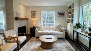 A living room with a fireplace, a sofa, armchairs, a coffee table, and a large window with a view of trees. - Charming Country Oasis (Alton)