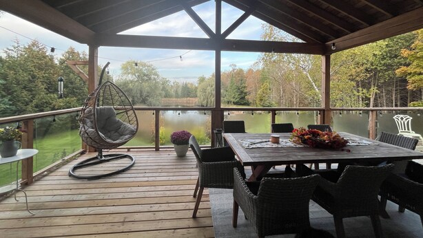 A covered deck with a wooden table and chairs, a hanging chair, and a view of a lake and trees. - Charming Country Oasis (Alton)