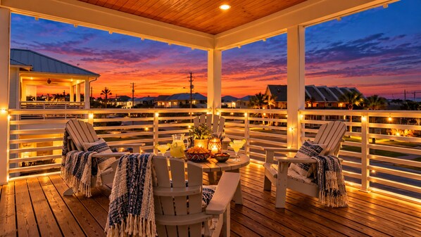 Un espacio de estar al aire libre con techo, con sillas y una mesa, con vistas a un barrio residencial al atardecer.