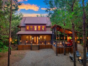 A two-story cabin with a porch, surrounded by trees and a gravel driveway.