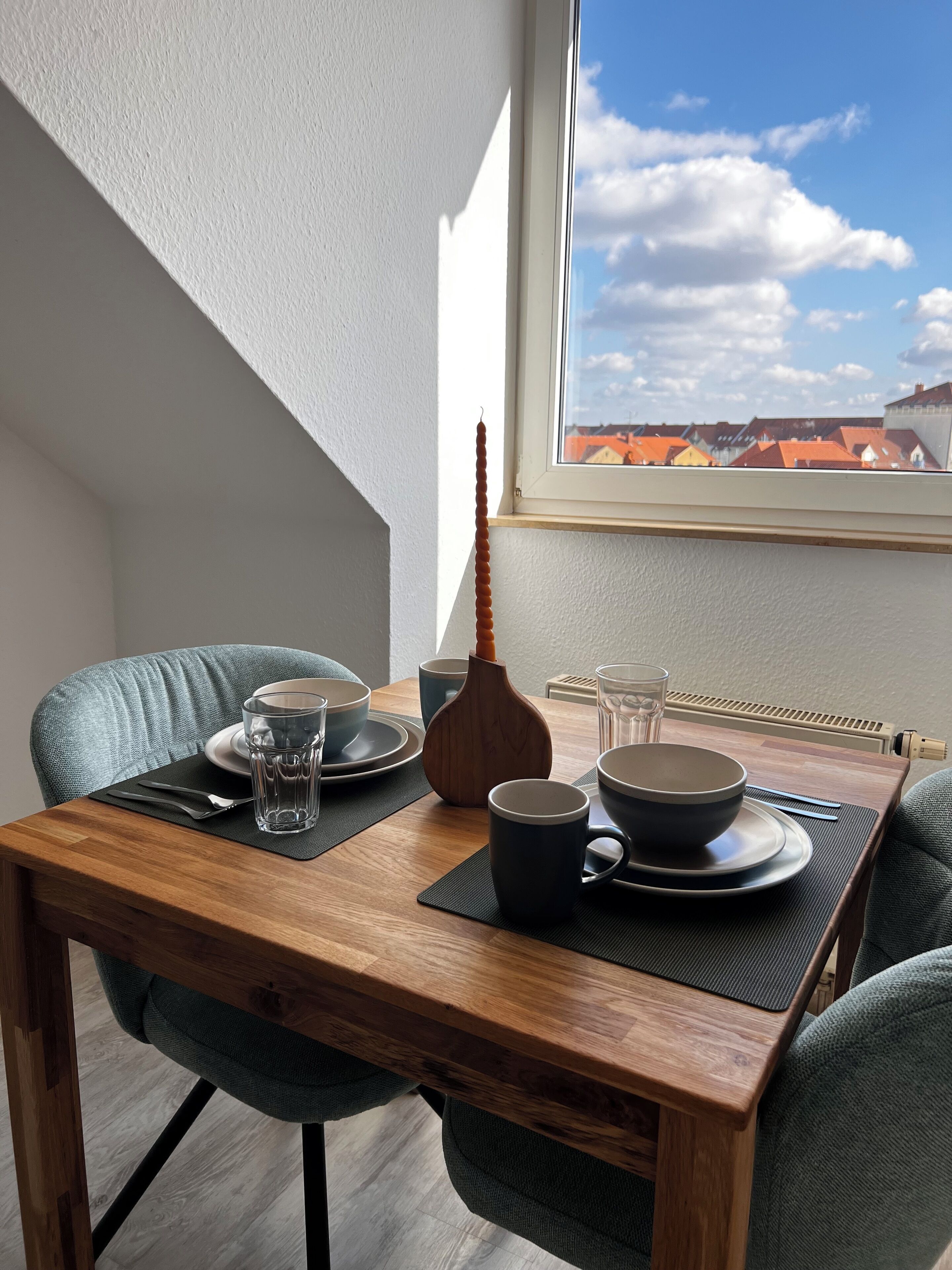A dining area with a wooden table, two chairs, a vase, and a view of buildings through a window.