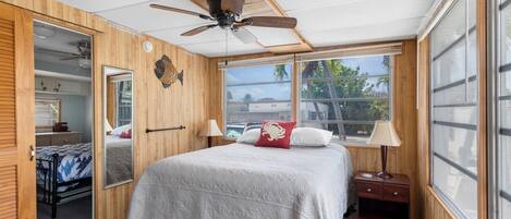 Une chambre à coucher comprenant un lit, un ventilateur de plafond, une table de chevet, une lampe, une fenêtre et une porte donnant sur une autre pièce.