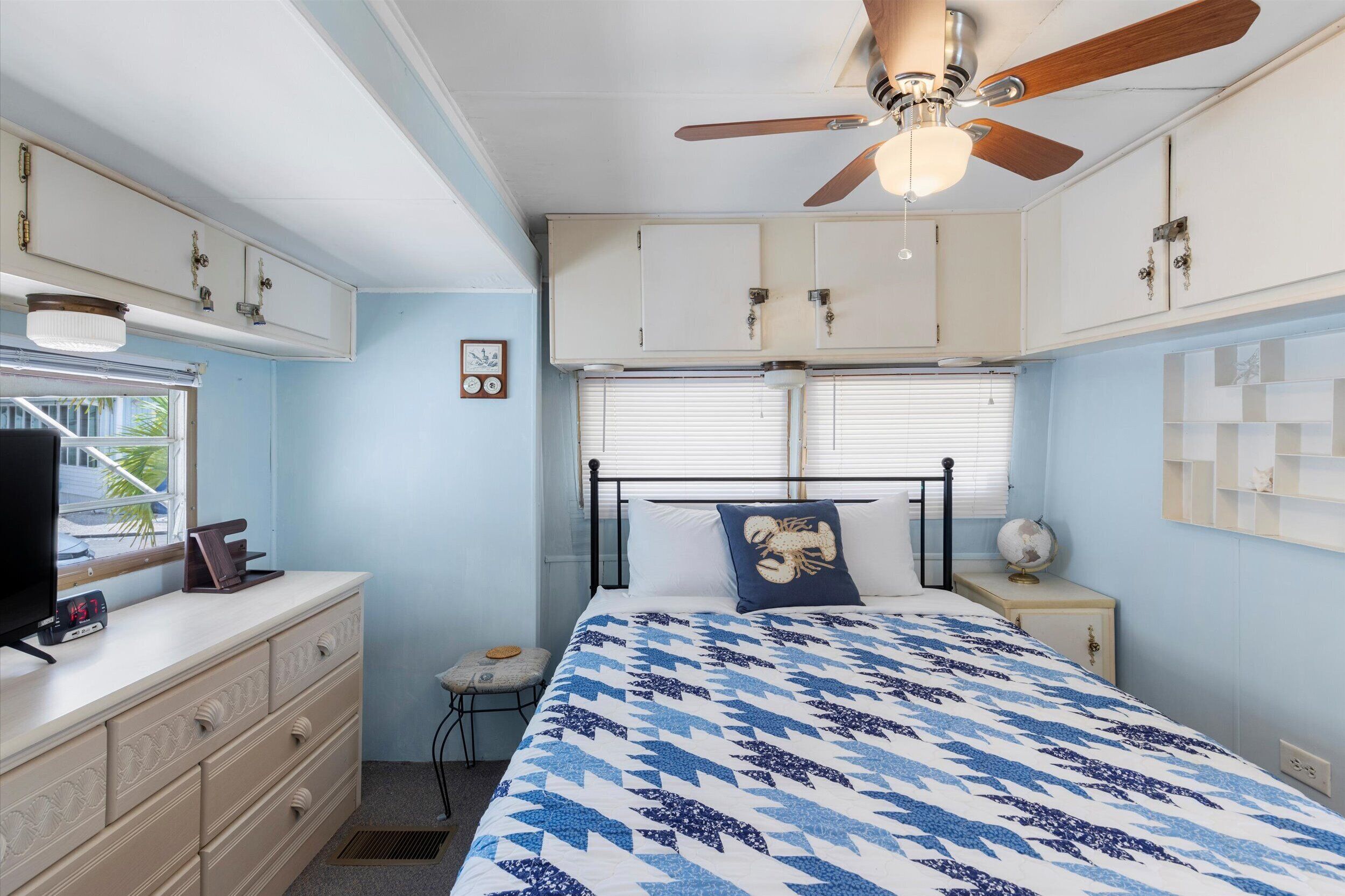 Une chambre à coucher comprenant un lit, un ventilateur de plafond, une commode, une table de chevet, une lampe et une fenêtre avec des stores.