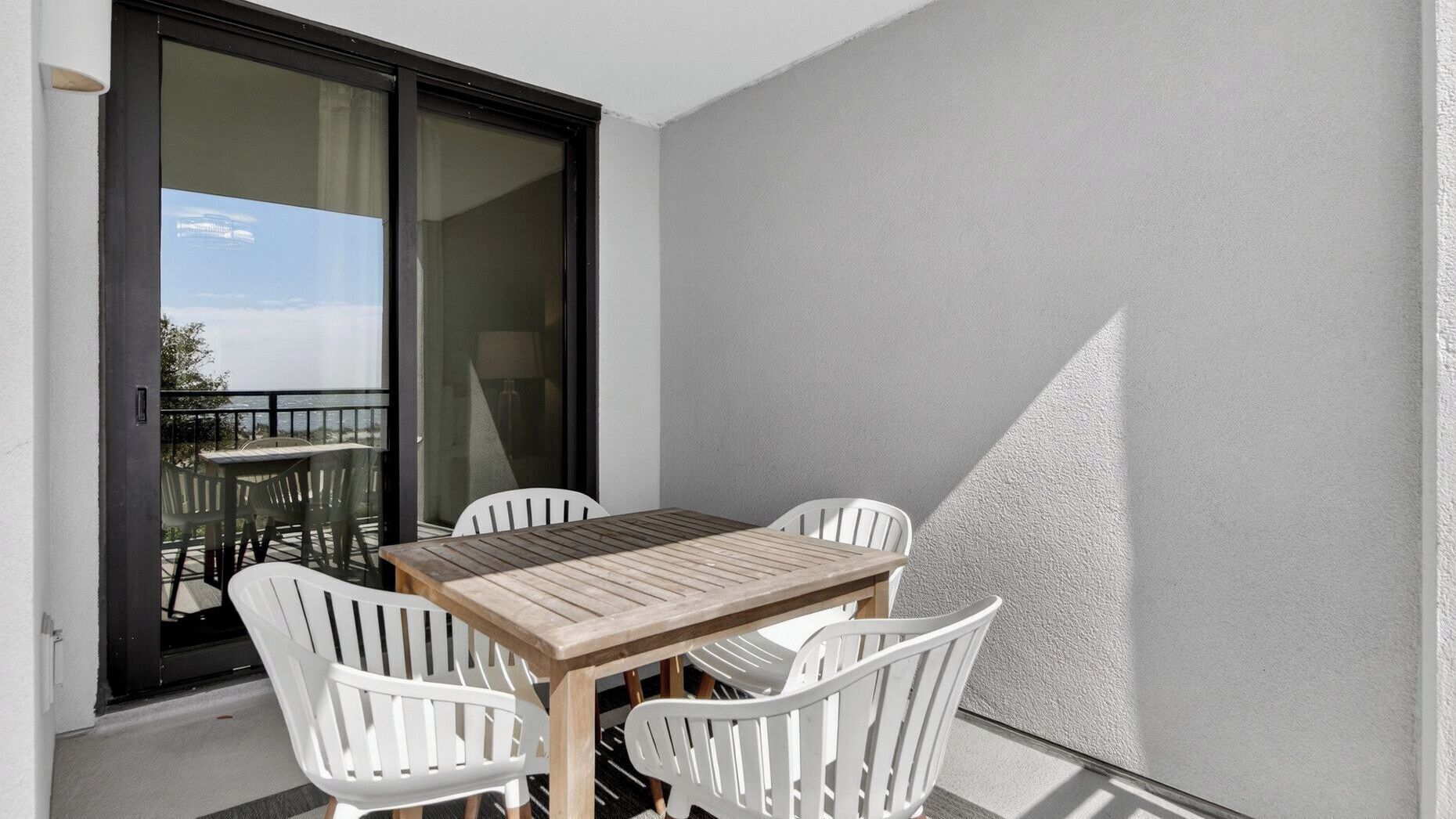 Un balcón con mesa de madera y sillas blancas, una puerta corredera de vidrio y vista al mar.
