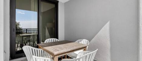 Un balcón con mesa de madera y sillas blancas, una puerta corredera de vidrio y vista al mar.