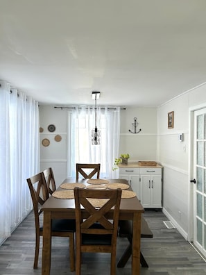 Uma sala de jantar com mesa e cadeiras de madeira, luminária pendente e cozinha com armários e janela.