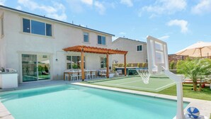 Ein Swimmingpool mit Basketballkorb, eine überdachte Terrasse und ein Haus mit Glasschiebeflügeln.