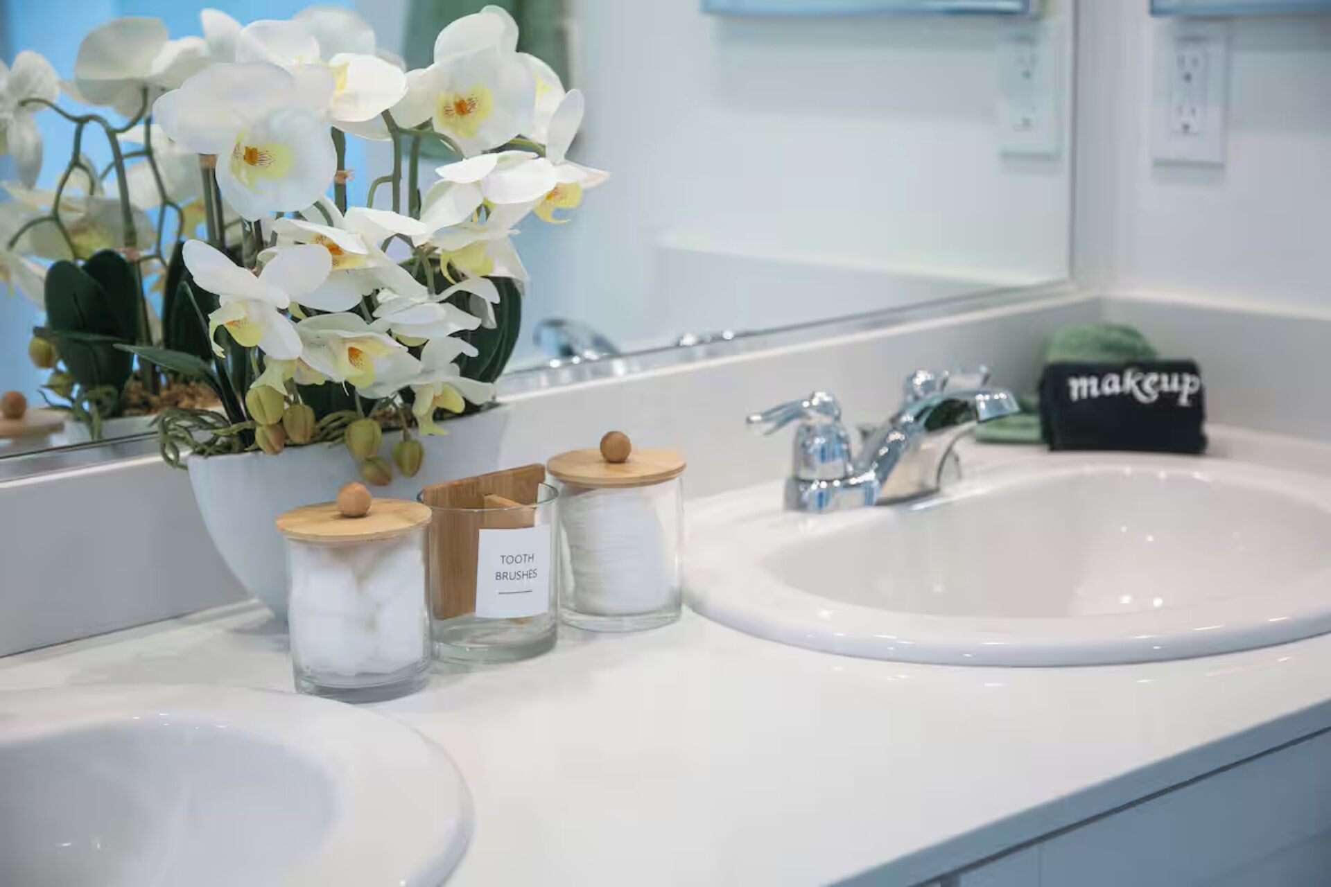 Un baño con un lavabo blanco, un espejo y un jarrón con flores blancas. Hay tres frascos con tapas de madera, uno etiquetado como 'TOOTH BRUSHES'.