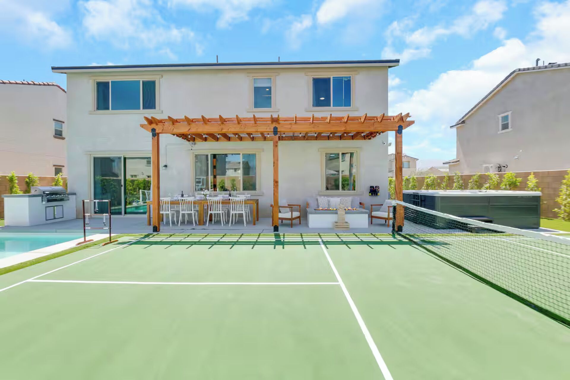 A tennis court with a wooden pergola and seating area, a pool, and a two-story building with large windows.