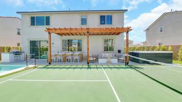 A tennis court with a wooden pergola and seating area, a pool, and a two-story building with large windows.
