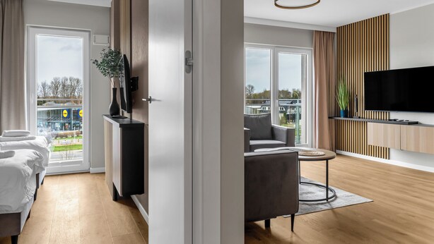 Une chambre d’hôtel moderne équipée d’un lit, d’un coin salon avec un fauteuil et une table, d’une télévision et d’une grande fenêtre donnant sur la ville.
