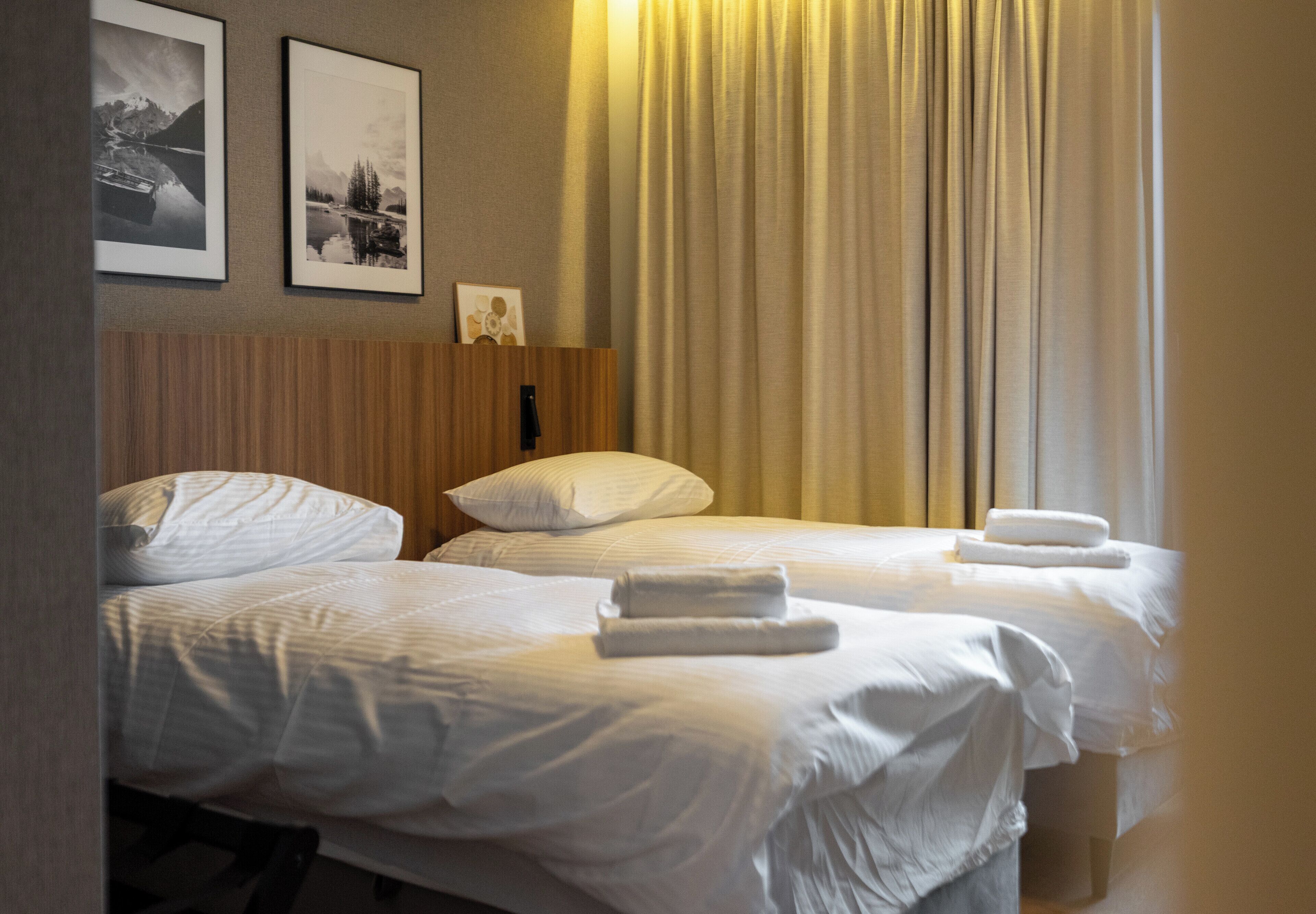 A hotel room with two beds, white linens, a wooden headboard, a nightstand, and a wall-mounted lamp.