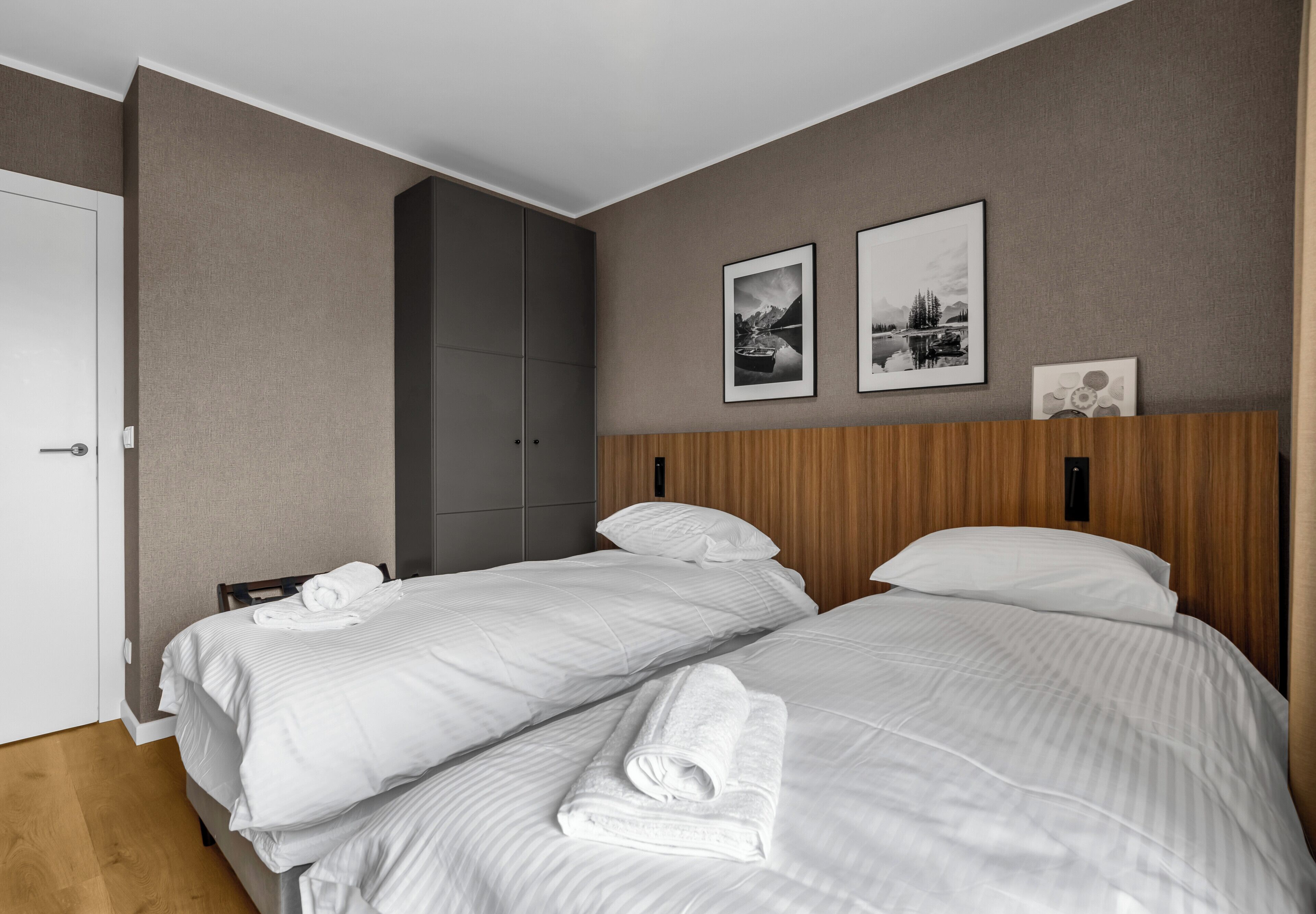 A hotel room with two beds, a wooden headboard, a wardrobe, and two framed pictures on the wall.